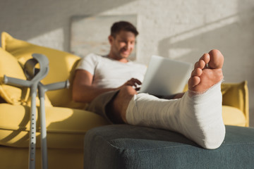 Selective focus of freelancer with broken leg on ottoman using laptop on couch