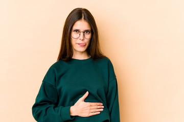 Young caucasian woman isolated on beige background touches tummy, smiles gently, eating and satisfaction concept.