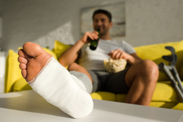 Selective focus of man with broken leg drinking beer and eating popcorn on couch in living room