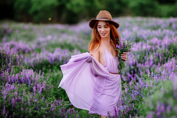 Red-haired girl in a hat lies in the grass with purple flowers. Young woman smile in nature. lady walks on a lavender field.