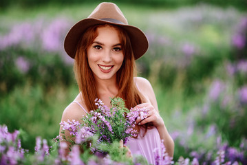 Red-haired girl in a hat lies in the grass with purple flowers. Young woman smile in nature. lady walks on a lavender field.
