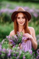Red-haired girl in a hat lies in the grass with purple flowers. Young woman smile in nature. lady walks on a lavender field.