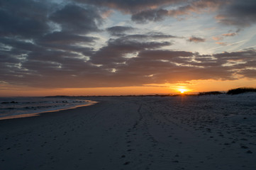 Fototapeta na wymiar The sun sets behind some clouds along the coastline of a sandy beach with a colorful sky.