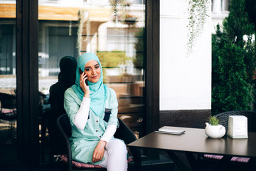 Beautiful young arabic girl in hijab talking by phone at cafe near the window.