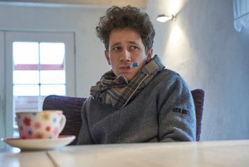 clouseup of young man sits at the table at home with a scarf around his neck and has a thermometer...