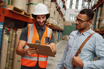 Storehouse factory worker discussing document with list of goods with warehouse manager holding...