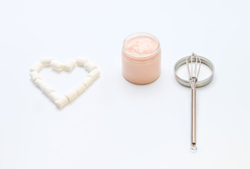 a glass transparent jar with a pastel peachy cream in an opened jar with a metal top and a clean metal whisk lying with a heart, concept of enjoying using and making handmade cosmetics