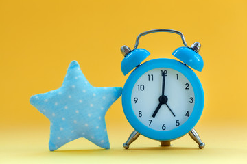 Soft blue pillow in shape of star and blue alarm clock on yellow background.