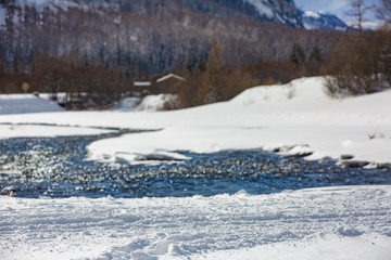 Snow trail in the foreground against the backdrop of a mountain river, alpine mountains and mountain hut. Selective focus, blur.