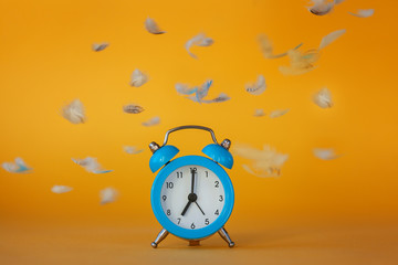 Soft fluffy flying feathers and blue alarm clock on yellow background.
