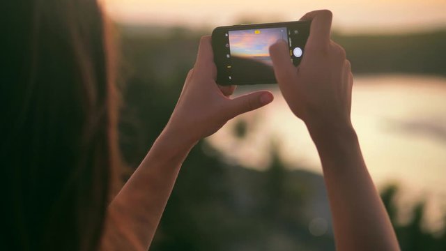 Close up rear view of young woman making photos and videos of beautiful sea sunset. Attractive female takes pictures standing on seashore at sundown. Woman taking pictures of beach at sunset 4k
