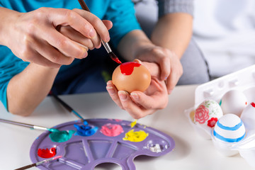 Fototapeta na wymiar Mother and her child hands painting Easter eggs. The mother helps her son paint an egg with a paintbrush at home. Happy family preparing for Easter. Easter holiday concept. Close up, selective focus