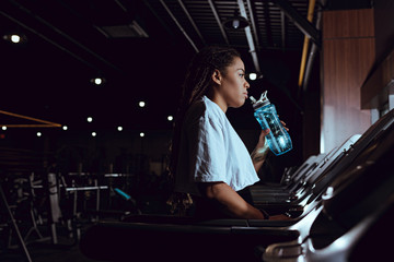 Fototapeta na wymiar Side view of african american woman drinking water from sports bottle on treadmill