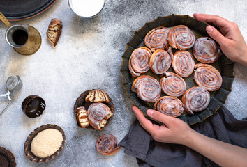 Cinnamon rolls or cinnabon with white cream glaze on a vintage plate, coffee, milk, brown sugar. Female hands holding a tray with fresh cinnabons. Gray background, top view, copy-space 