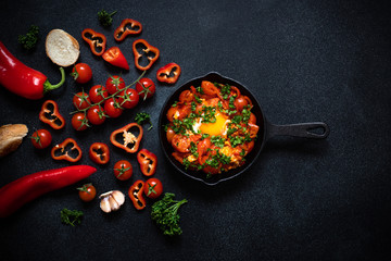 Obraz na płótnie Canvas Shakshouka eggs poached in a sauce of tomatoes, vegetables, spices and herbs in iron pan. Bread, tomatoes, red peppers, garlic, parsley. Traditional breakfast. Black background, top view, copy space
