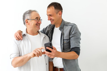 Obraz premium Happy two age generations men family old father embracing young grown adult son having fun enjoying using smart phone bonding watching funny social media video using mobile apps
