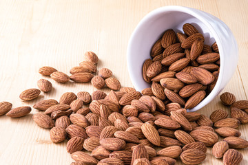 Closeup almonds nut are poured from white bowl on wooden table.