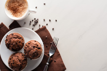 top view of fresh chocolate muffins on white plate and brown napkin near fork and cappuccino on...