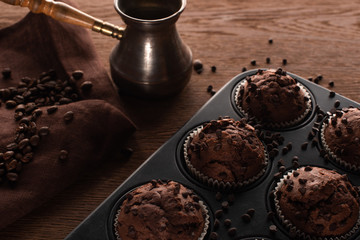 fresh chocolate muffins in muffin tin on wooden surface near cezve with coffee beans on napkin