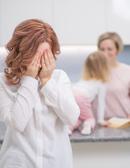 Obraz premium A teenage girl looks upset at the camera, in the background her mother is playing with her younger sister. Problems of puberty, depression, problems in the family