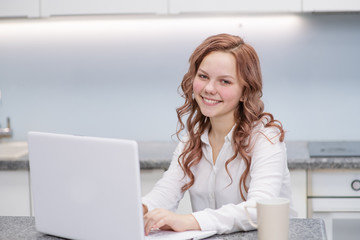 Teen girl working on a laptop while sitting at home in the kitchen, smiling while looking at the camera