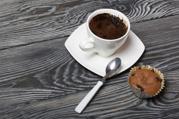 Coffee in a white cup stands on a saucer. Nearby lies a muffin, and a spoon on a saucer. On brushed pine boards.
