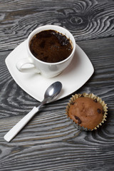 Coffee in a white cup stands on a saucer. Nearby lies a muffin, and a spoon on a saucer. On brushed pine boards.