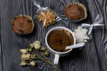 Coffee in a white cup. Nearby are muffins, and a dried rose of beige color. Sachets of cookies in the shape of snowflakes are visible. On brushed pine boards.