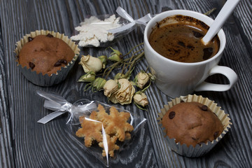 Coffee in a white cup. Nearby are muffins, and a dried rose of beige color. Sachets of cookies in the shape of snowflakes are visible. On brushed pine boards.