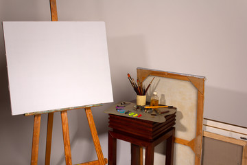 Oil painting. Blank canvas on Easel. Palette with paints and brushes. Squeeze out oil paints on a palette. Clean cloth with copy space. Set of brushes in a cup. Subframes and drawing tools.