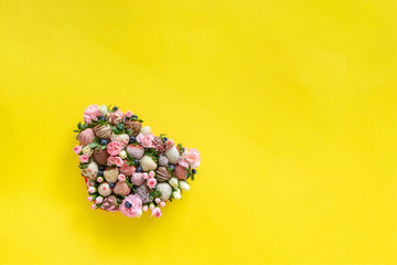 Heart shaped box with handmade chocolate covered strawberries with different toppings and flowers as a present on Valentines day on yellow background with free space for text - obrazy, fototapety, plakaty