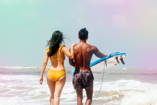 Back side of Young man holding serf board in hand and woman wearing yellow swimsuit walking on the beach .prepare for doing water activity,blurry light around