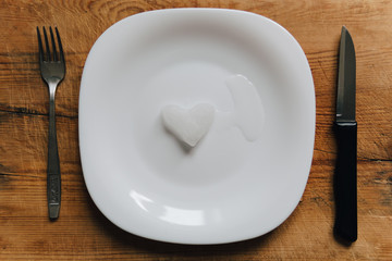 A snowy heart melts on a white plate. Heart shaped snow on a saucer. Ice in frosty weather. Melted water is collected in a small puddle. Wooden background. Love on valentine's day concept