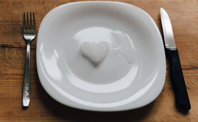 A snowy heart melts on a white plate. Heart shaped snow on a saucer. Ice in frosty weather. Melted water is collected in a small puddle. Wooden background. Love on valentine's day concept