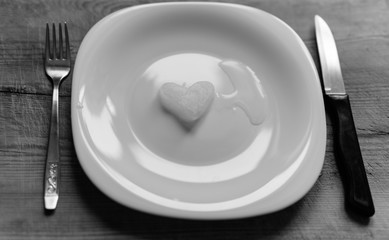 A snowy heart melts on a white plate. Heart shaped snow on a saucer. Ice in frosty weather. Melted water is collected in a small puddle. Wooden background. Love on valentine's day concept