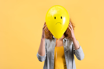 young woman holding sad balloon in front of face, isolated on yellow