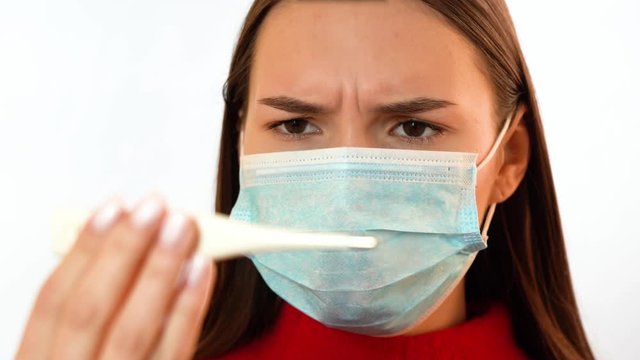 A girl in a medical mask looks at a thermometer and frowns.