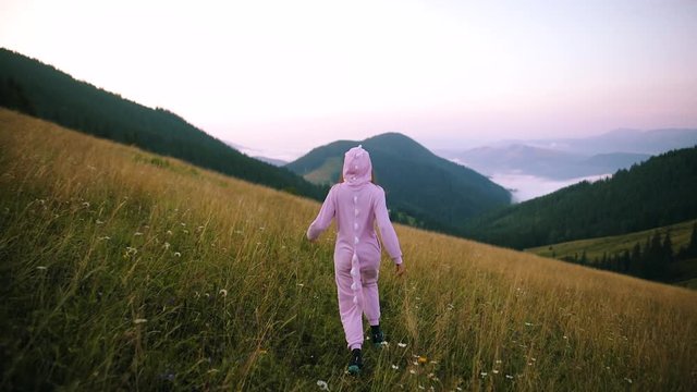 Slim positive young woman in pink night-suit running on green grass on top of a mountain. Happy girl having fun in nature on summer meadow. Woman hiker in pink Dragon Pajamas