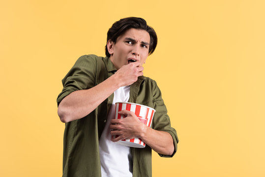 Frightened Young Man Watching Horror Movie And Eating Popcorn, Isolated On Yellow