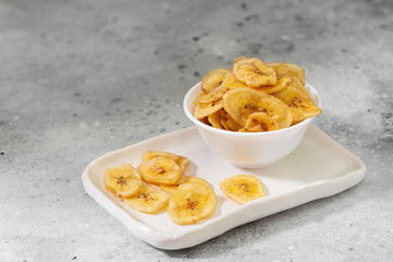 Dried bananas in a white ceramic bowl on a gray kitchen table. Banana chips. Vegetarian snack for proper nutrition