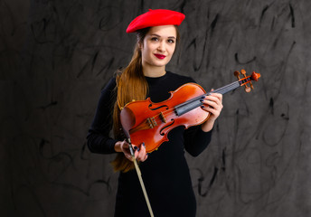 Portrait of a pretty brunette musician girl in a black dress and red beret on a gray background plays the violin