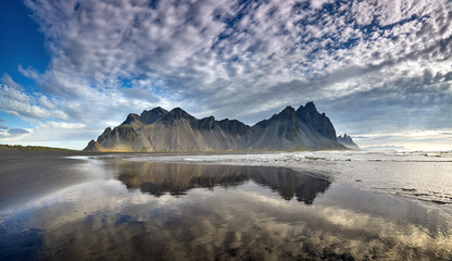 Panorama of Famous Stokksnes mountain reflected in water on Vestrahorn cape (Batman Mountain), Iceland, Europe. Popular tourist attraction. Beauty world. Black sand dunes on the beach. Postcard.