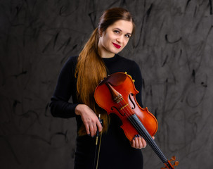 Portrait of a pretty brunette musician girl in a black dress on a gray background holds a violin in her hands