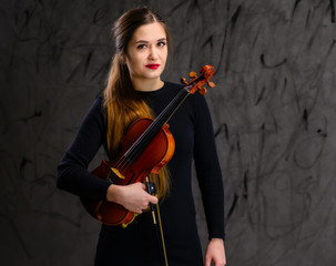 Portrait of a pretty brunette musician girl in a black dress on a gray background holds a violin in...