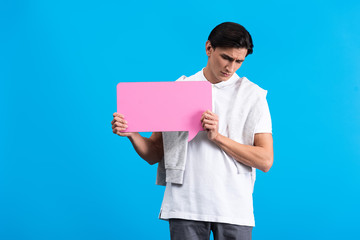 upset young man holding pink speech bubble, isolated on blue