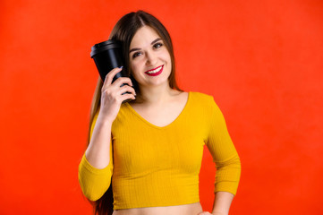Obraz premium nice brunette girl with long hair with a smile with a cup of coffee in her hands in a yellow sweater is happy over a red background smiling and showing positive emotions