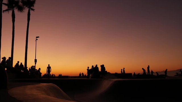 Silhouette of young jumping skateboarder riding longboard, summer sunset background. Venice Ocean Beach skatepark, Los Angeles California. Teens on skateboard ramp, extreme park. Group of teenagers