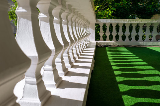 Semi-abstract of a white stone pillar wall with leading lines and regular patterns of light and shadow cast on a green floor by strong directional sunlight.