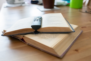 Close-up of two open books, one on top of the other, with a television remote control on the top, all set on a table with shallow depth of field and soft-focus items in the background.