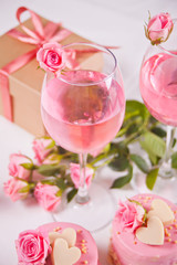 Obraz na płótnie Canvas Two glasses with pink grape wine with rose flowers and mini cakes. Romantic dinner concept.
